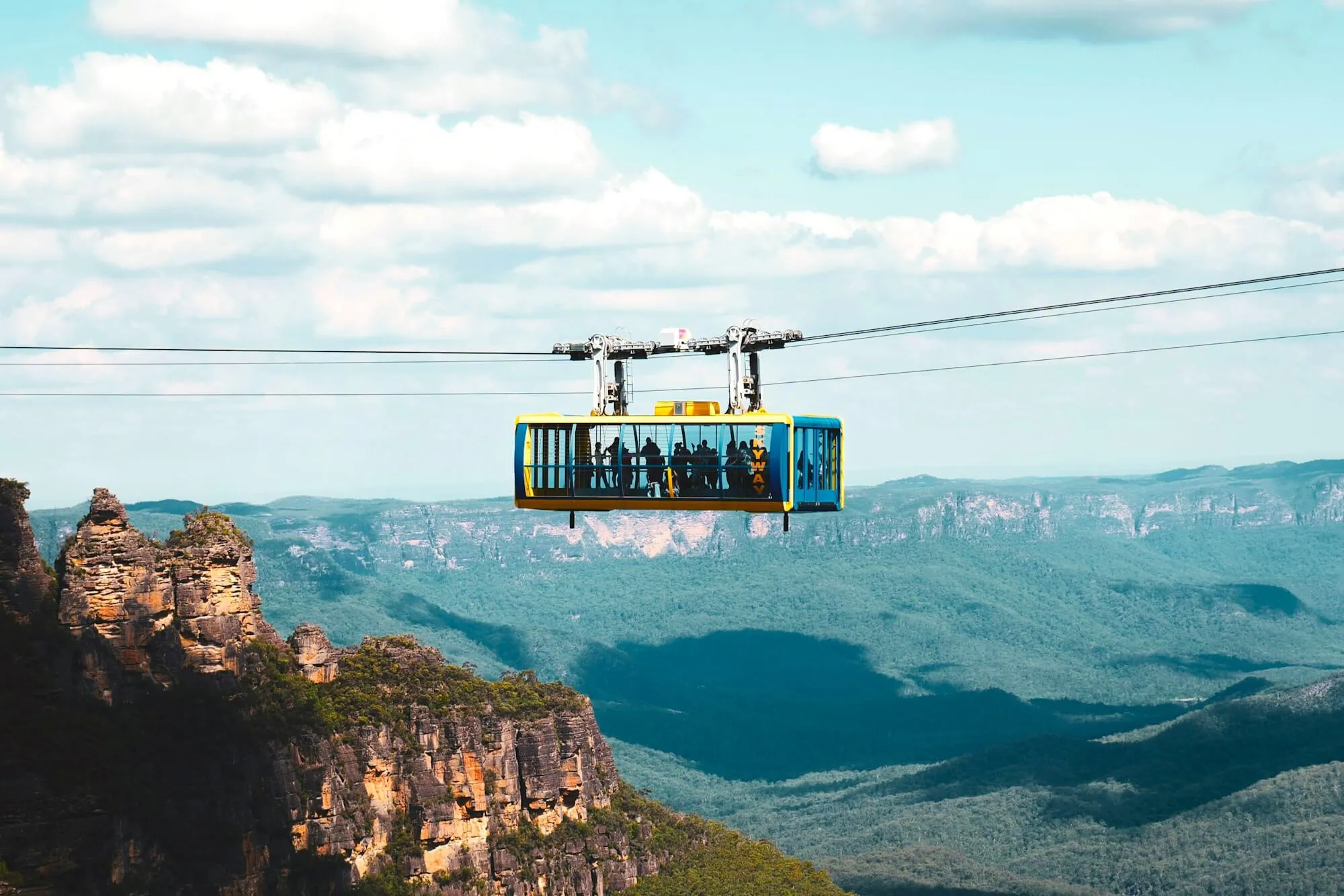 Yellow-and-blue Skyway cabin full of riders crossing above the Jamison Valley, the Three Sisters below on the left